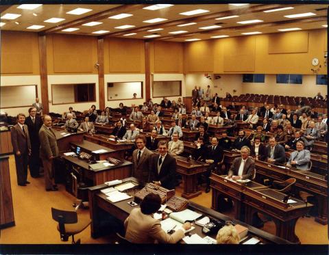 Color photo of the Assembly in session in the Temporary Chamber, 1979.  The Assembly met in these temporary chambers in Capitol Park during the Capitol restoration, 1976-1981. Photo shows Members seated at their desks on the Floor. Standing at dais: Speaker Leo McCarthy, Speaker pro Tempore John T. Knox, and Assistant Speaker pro Tempore Tom Bane.