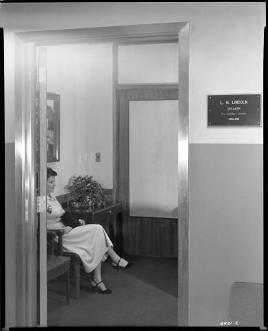 Photo looking in through the entrance door of the Office of Speaker Luther Lincoln (R-Oakland), circa 1955. Photo shows woman visitor seated in the reception area with purse on lap (black and white photo).