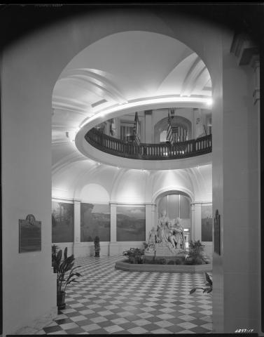 Photo taken from public entrance on first floor rotunda looking toward the Columbus/Queen Isabella statue.  Visible behind the statue is the newly constructed Annex hallway.  On the left wall is a large bronze plaque entitled “Logan’s Memorial Day Order.”  Large murals painted by Arthur Matthews from the Bay Area (mounted in 1915) are on the rotunda walls depicting California historical scenes. These murals were moved to the basement rotunda during the 1976-81 restoration. (black and white photo).