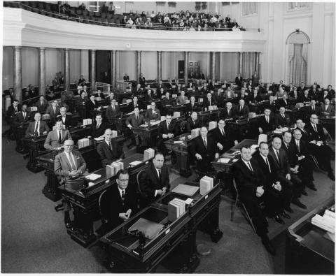 Assembly in session, circa 1963.  Photo shows members seated at their desks.  Speaker Unruh and Assembly Members Knox, Marks, Garrigus, Mills, and others seated in front on folding chairs.