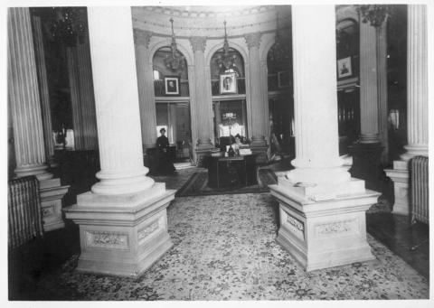 Interior view of State Library housed in the Capitol apse upper level, circa 1905. Female librarians pictured wearing Victorian era clothing. Paintings of Shakespeare and other literary figures hanging on wall (black and white photo).