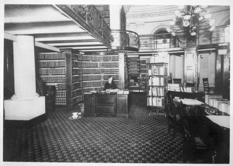 Interior view of State Library housed in the Capitol apse lower level, circa 1905. Male librarian pictured sitting at desk in area of the book “stacks.” Gas chandelier and ornate iron railings shown (black and white photo).