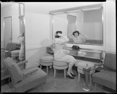 Ladies “powder room” in newly constructed Capitol Annex in 1952. Woman shown looking into mirror while seated in powder room, with purse on shelf. 1950s era furniture (black and white photo).