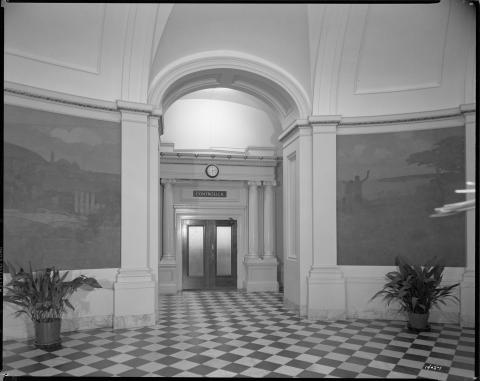 Interior view of the first floor State Capitol rotunda (circa 1952), showing entrance to the State Controller’s office.  Large murals painted by Arthur Matthews from the Bay Area (mounted in 1915) are on the rotunda walls depicting California historical scenes. These murals were moved to the basement rotunda during the 1976-81 restoration. Checker pattern floor tile shown. Plain white paint shown on rotunda ceiling arches, illustrating how utilitarian the rotunda had become by the mid-20th Century (black an