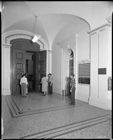 View of the hallway outside the main Senate Chamber entrance, circa 1952. State Police officer holding door open to admit citizens.  Committee bulletin board on wall. Sign on wall directing visitors to public gallery on the Third Floor. Note the fluorescent lighting fixture (black and white photo).