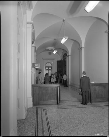 View of the hallway outside the main Assembly Chamber entrance, circa 1952. State Police officer holding door open as visitors peer into Chamber. Across from the Chamber doors note the press offices:  Oakland Tribune, Fresno and Modesto Bee, SF and LA Examiner,  and SF Call-Bulletin (black and white photo).