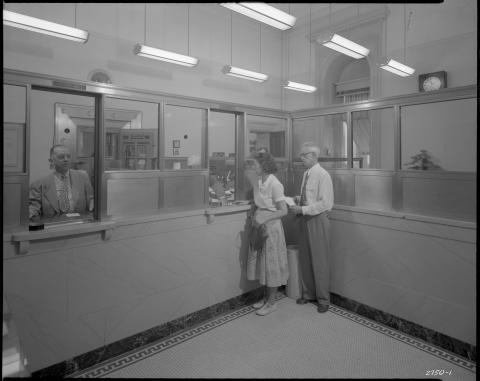 View of the State Treasurer’s office on first floor of the West Wing of the Capitol, August 19, 1952 (per the wall calendar). Security glass sits atop long public counter, with slots to slide paperwork, checks, and cash.  A large metal vault door is seen behind counter.  “Receiving” window is seen on the right (with two persons lined up to do business with a Treasurer staff person); and “Paying” window on the left, showing another worker at his post (black and white photo).