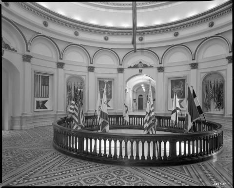 1950s photo of second floor rotunda, looking south toward the Senate.  Several flags are shown mounted on the rotunda railing. Additional flags are displayed in cases in the rotunda wall niches. Visible on the Senate side of the rotunda are signs reading: “Women’s Restroom;” “Pacific Telephone Public Telephones;” “Lieutenant Governor;” and “Legislative Bill Room.” (black and white photo).