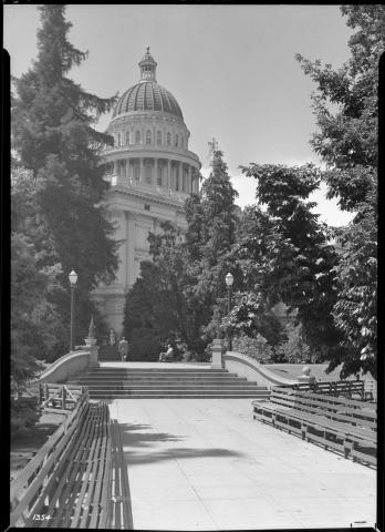 Pre-Annex era photo:  exterior view of Capitol building from northeast looking toward Assembly side of building.  Terraced landscape is visible in Capitol Park, with steps connecting terrace segments. Numerous park benches and trees are also visible.  Man seated on park bench. Man and woman seen walking near Capitol (black and white photo).