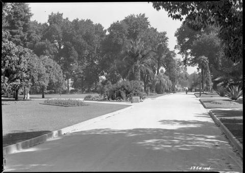 View of eastern Capitol Park looking eastward on center walking path toward 15th Street, circa 1948.  Visible is a small sundial mounted on a stone; people walking through the park; running sprinklers; and a wooden tower structure on wheels in the distance, near 15th street (black and white photo).