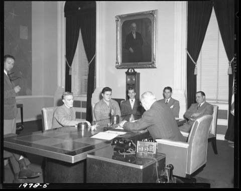 Photo of Governor Earl Warren meeting with 6 adults in his office, circa 1948.  Governor is seen seated at his large desk, with five men seated across from him, and one man standing.  Visible is a 1940s era phone and intercom system to the Governor’s left. Hanging on the wall is a portrait of California’s first Governor, Peter Burnett. Two small elephant figurines are on the desk. Desk appears to have checkered pattern inlay. Grandfather clock is stationed beneath Burnett portrait.