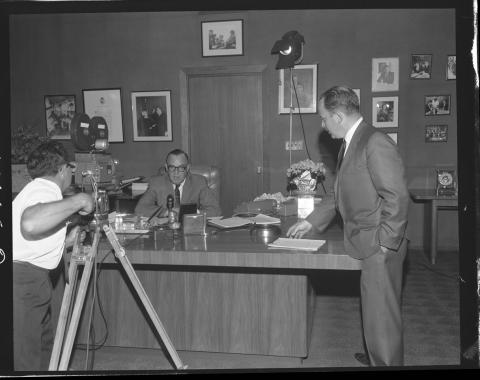 Image of Governor Edmund G. “Pat” Brown, sitting at his desk during what appears to be a televised address, circa early 1960s.  Film crew with tripod and camera is visible.